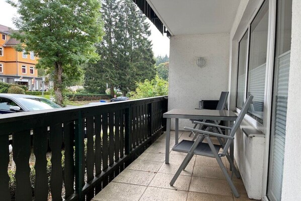 Ein Balkon mit Tisch und Stühlen, einem schwarzen Geländer und Blick auf geparkte Autos und Bäume.