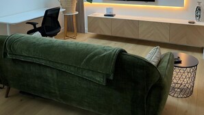 Ein modernes Wohnzimmer mit einem an der Wand montierten Flachbildfernseher, einem Schreibtisch mit Stuhl, einem grünen Sofa und einem kleinen Beistelltisch mit einer Dekoration.
