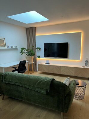 A modern living room with a flat-screen TV mounted on the wall, a desk with a chair, a green sofa, and a small side table with a decorative item. - The Wing is a luxury and peaceful private retreat for two adults. (New Milton)