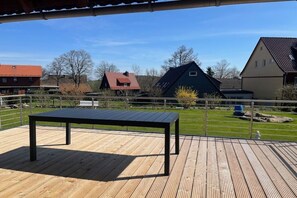Een houten terras met een tafel, omringd door een metalen hek, met uitzicht op een woonwijk met huizen en een heldere lucht.