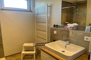Een badkamer met een wit wastafelmeubel, een douche, een handdoekrek en een klein opstapkrukje.