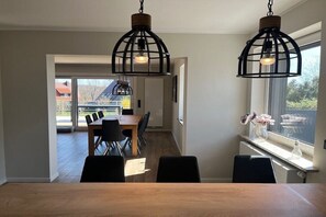 Een eetgedeelte met een houten tafel en stoelen, twee hangende hanglampen en een uitzicht door een raam.