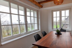 Un espace repas avec une table et des chaises en bois, de grandes fenêtres donnant sur des arbres et un bâtiment, et un plafond avec des poutres apparentes.