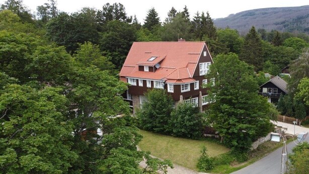 Uma casa de dois andares com telhado vermelho, cercada por árvores verdes e densas.