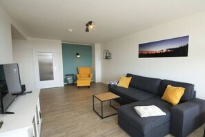 Een woonkamer met een donkere bank, een gele fauteuil, een salontafel en een televisie op een meubel.