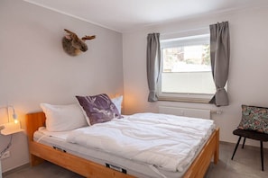 Une chambre à coucher avec un lit, une décoration murale représentant une tête d’animal empaillée, une fenêtre avec des rideaux et une chaise avec un coussin.