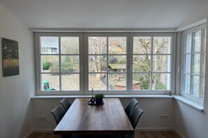Un coin repas avec une table et des chaises en bois, de grandes fenêtres donnant sur l’extérieur et une petite plante posée sur la table.