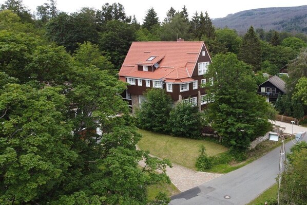 Une maison à deux étages, avec un toit rouge, entourée d’arbres verdoyants et touffus.