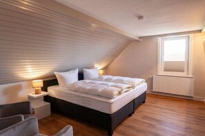 Une chambre avec un plafond en pente, un lit avec une literie blanche, une table de chevet avec une lampe et une fenêtre avec des stores.