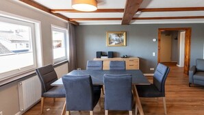 Quarto com teto de madeira, uma mesa com cadeiras, radiador e janela com vista para edifícios.