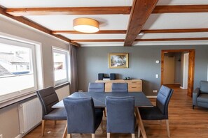 Une pièce avec un plafond en bois, une table entourée de chaises, un radiateur et une fenêtre donnant sur des immeubles.