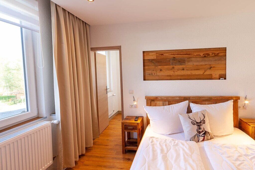 A bedroom with a wooden headboard, white bedding, a deer pillow, and a view through a window with curtains.
