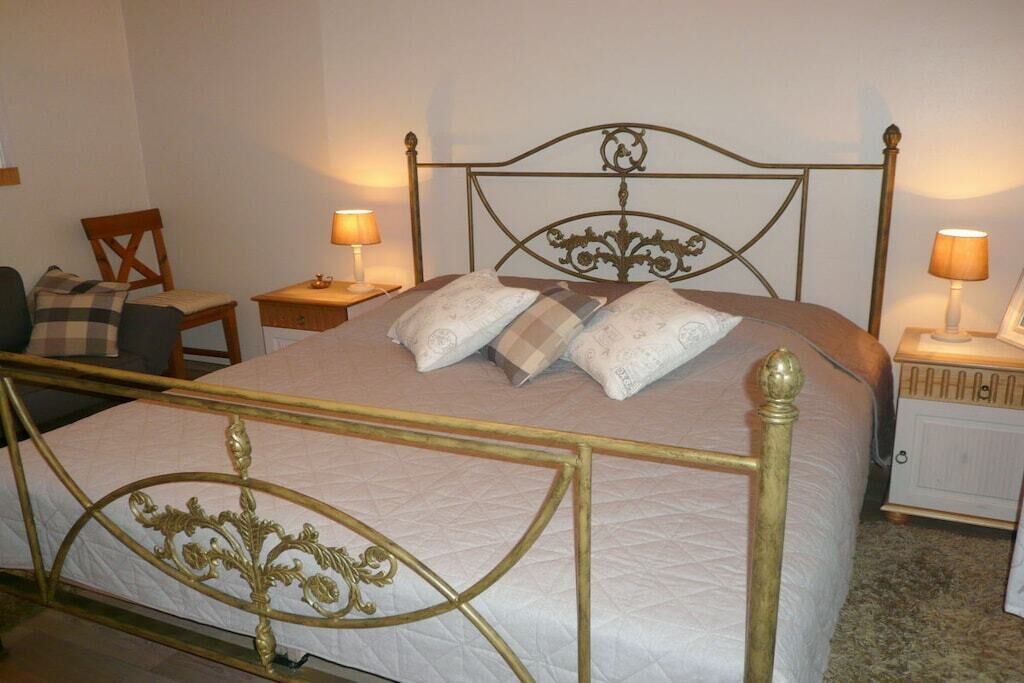 Uma cama com estrutura metálica e cabeceira, duas mesas de cabeceira com abajures, uma cadeira e um pequeno armário.