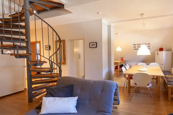 Ein modernes Wohnzimmer mit einer Wendeltreppe, einem grauen Sofa, einem Esstisch mit Stühlen und einer Wand mit einem Motivationsspruch.