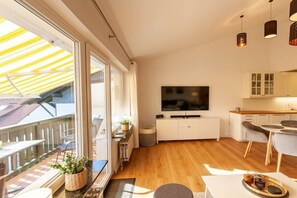 A modern living room with a flat-screen TV, wooden flooring, a balcony with a yellow awning, and a kitchen area with white cabinets. - Eye-catching with a dream view (Braunlage)