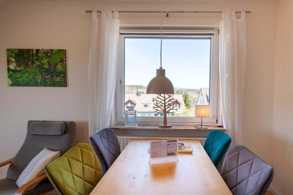 A room with a dining table, chairs, a pendant light, a window with curtains, and a painting on the wall.
