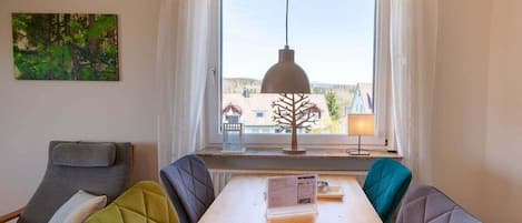 A room with a dining table, chairs, a pendant light, a window with curtains, and a painting on the wall.