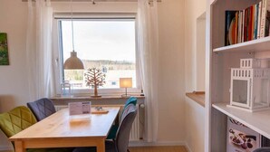 Uma sala de jantar com mesa e cadeiras de madeira, uma janela com cortinas, uma luminária pendente e uma estante com livros e uma taça de vinho.