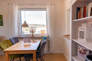 Un coin repas avec une table et des chaises en bois, une fenêtre avec des rideaux, un luminaire suspendu et une bibliothèque contenant des livres et un verre à vin.