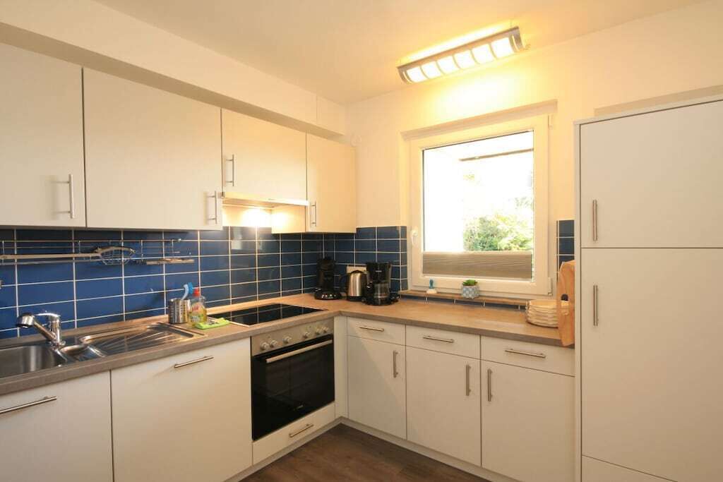 Una cocina compacta con armarios blancos, horno empotrado, lavamanos y ventana con persianas.