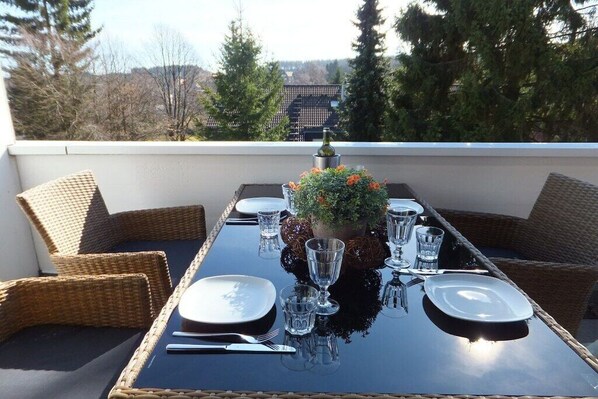 Une table à manger dressée pour quatre personnes sur un balcon, avec des chaises en osier, une plante en pot et une bouteille de vin.