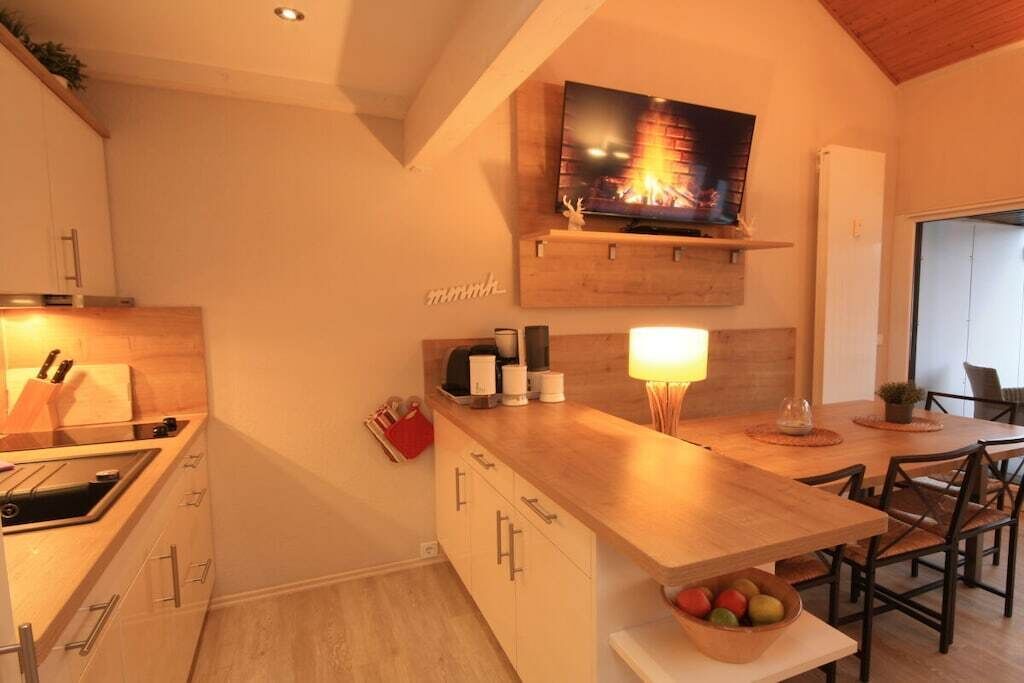A modern kitchen with a built-in stove, wooden cabinets, a dining area with a wooden table and chairs, and a mounted television above the stove.