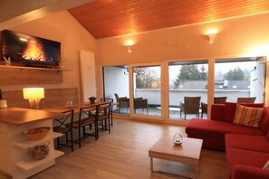 Un salon avec un téléviseur, un coin repas avec des chaises, un canapé rouge et une porte-fenêtre coulissante donnant sur une terrasse extérieure.