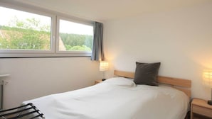 Quarto com cama, mesa de cabeceira, luminária, janela e vista para o verde lá fora.