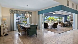 Ampla sala de jantar com uma mesa grande e cadeiras, um lustre e vista para um pátio externo através das portas de vidro.