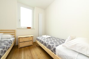Ein kleiner, sauberer Raum mit zwei Holzbetten, einem Nachttisch aus Holz und einem Fenster, das natürliches Licht hereinlässt.