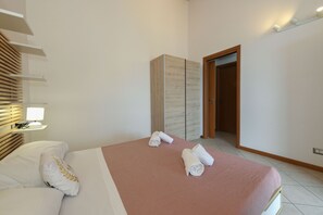 A hotel room with a bed, a nightstand, a lamp, a wooden wardrobe, and a door leading to another room. - MGH - Tavaredo Lake Retreat, Polpenazze (Polpenazze del Garda)