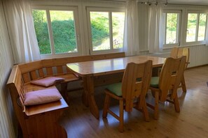 A room with a wooden dining table and chairs, a bench with cushions, and large windows with a view of greenery. - Holiday apartment with 1 bedroom (Kleines Wiesental - Neuenweg)