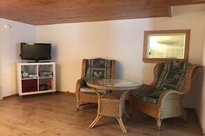 A room with a television on a white stand, two wicker chairs with cushions, a small round table, and a window with a mirror. - Holiday apartment with 1 bedroom (Kleines Wiesental - Neuenweg)