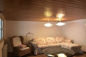 Une pièce avec un plafond en bois, un canapé à motifs floraux, un fauteuil en osier et une table basse ronde en verre.