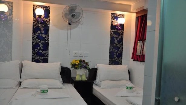 Habitación de hotel con dos camas individuales, una mesita de noche con un jarrón de flores, un ventilador de pared y una cortina roja.