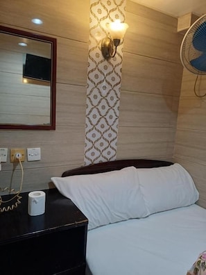 A hotel room with a bed, a bedside table with a lamp, a wall-mounted fan, and a mirror. - Everest Base Camp (Kowloon)