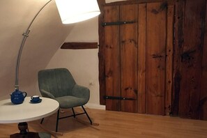 Een kamer met een houten vloer, een ronde tafel met een blauwe theepot en kopje, een groene fauteuil en een houten deur met metalen scharnieren.