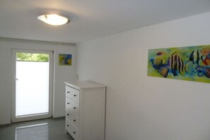 Une pièce avec une commode blanche, une porte donnant sur l’extérieur et un tableau coloré accroché au mur.