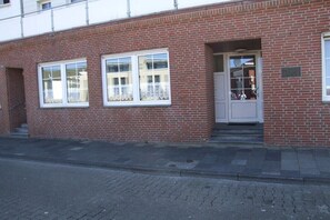 A red brick building with two windows and a door, a paved sidewalk, and a metal fence. - Holiday apartment with 1 bedroom (Norderney)