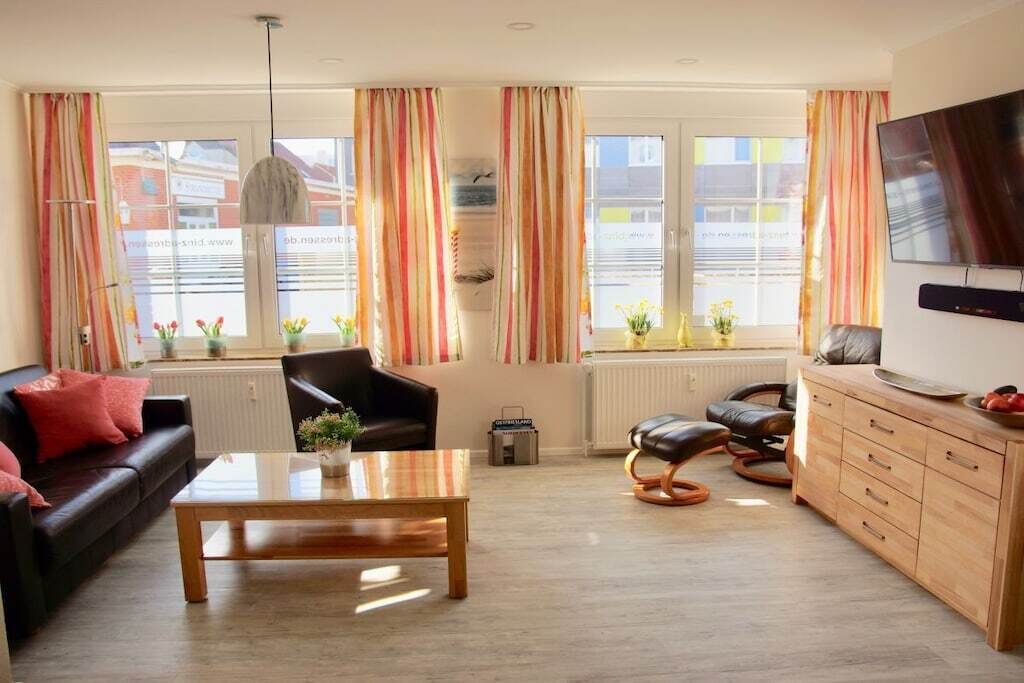 A living room with a sofa, coffee table, and TV. There are windows with curtains, a radiator, and a plant.
