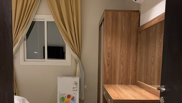 A compact kitchen with a refrigerator, wooden cabinets, and a window with curtains. - Alamaan Servised Apartment  (Madinah)