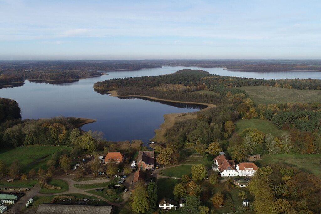 Une propriété située au bord d’un lac, comprenant plusieurs bâtiments, dont une grande maison blanche avec un toit rouge, entourée d’arbres et de vastes champs.
