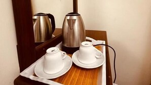 Una mesa de madera con una bandeja que sostiene dos tazas y platillos blancos, un hervidor eléctrico plateado y un enchufe.