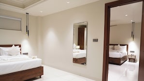 A hotel bedroom with a large bed, a bedside table, a nightstand, a mirror, and a door leading to another room. - Kashi Courtyard (Varanasi)