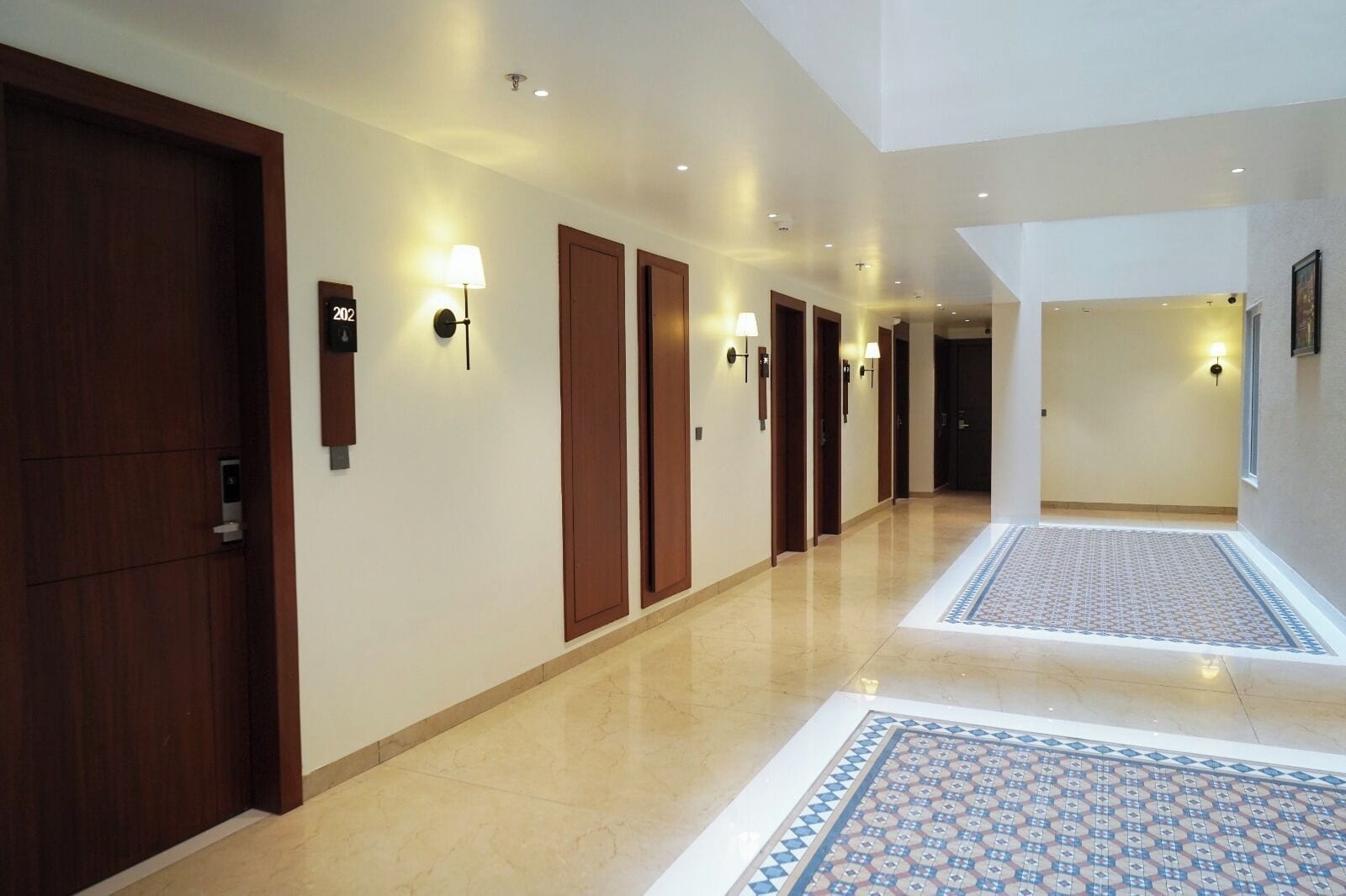 Un pasillo de hotel con varias puertas, luminarias fijadas a la pared y alfombras estampadas en el suelo.