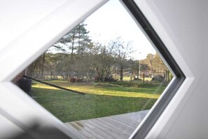Une vue à travers une fenêtre donnant sur une terrasse en bois, une pelouse et des arbres en arrière-plan.