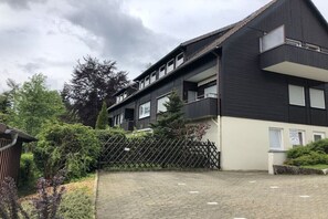 A multi-story residential building with a dark exterior, a paved driveway, and a wooden fence. - Holiday apartment with 1 bedroom (Hahnenklee Bockswiese)