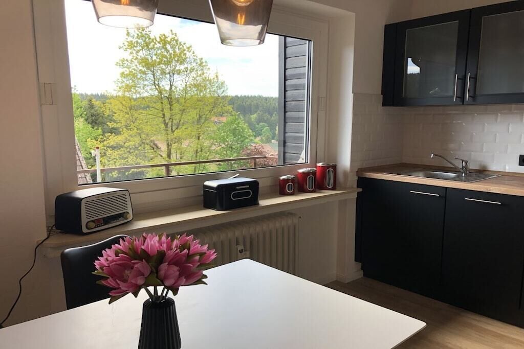 Una cocina con ventana que da a un bosque, una mesa blanca con un jarrón de flores y armarios negros.
