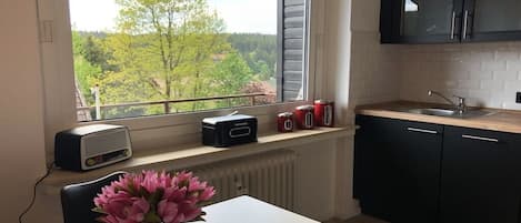 Una cocina con ventana que da a un bosque, una mesa blanca con un jarrón de flores y armarios negros.