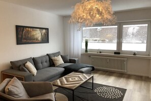 Ein modernes Wohnzimmer mit einem grauen Ecksofa, einem Couchtisch mit geometrischem Muster und einem großen Fenster mit Blick auf ein Gebäude.